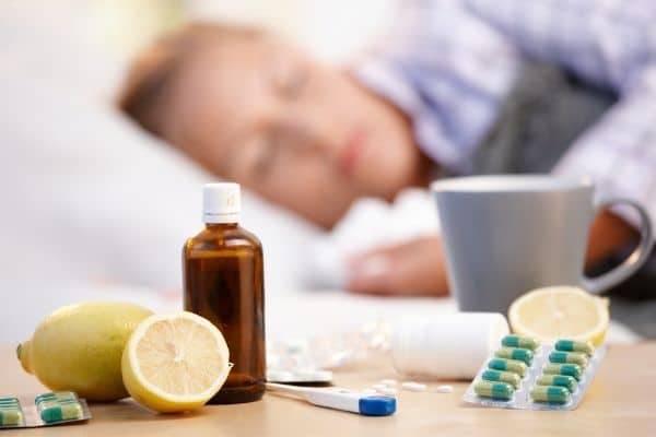 A wide variety of OTC cough and cold medicine on a bed side table as a woman sleeps in the background